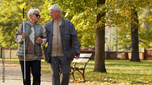 Senior Caucasian blind lady with glasses and a stick relies on her husband while having a walk in the park disabled blind people support concept medium full shot copy space. High quality photo