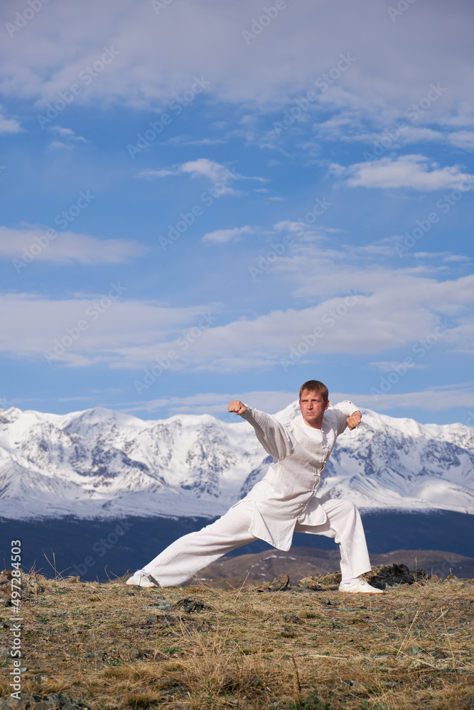 Wushu master in a white sports uniform training kungfu in nature on background of snowy mountains. Wushu master in a white sports uniform training kungfu in nature on background of snowy mountains.
