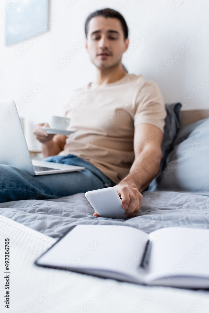blurred man holding cup of coffee while reaching smartphone in bedroom. blurred man holding cup of coffee while reaching smartphone in bedroom.