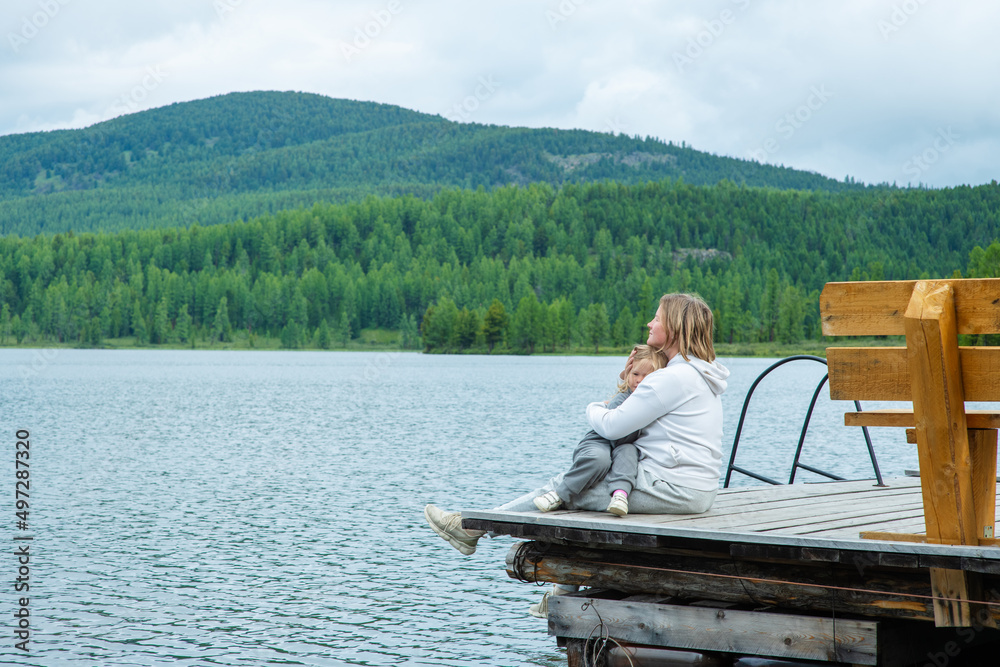 Mom hugs the child sitting next to her on the pier of a blue lake in the mountains and gently looks at the child. Family tourism