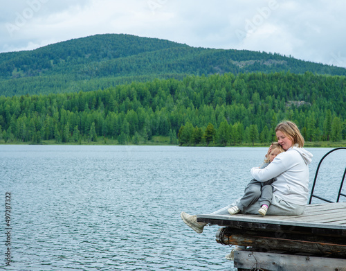 Mom hugs the child sitting next to her on the pier of a blue lake in the mountains and gently looks at the child. Family tourism