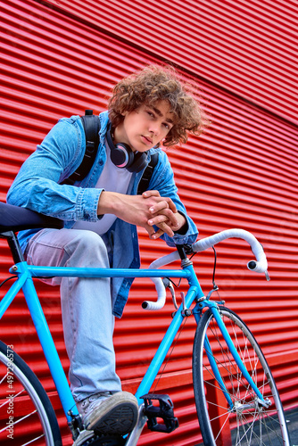 Low angle of curly haired teenage male with headphones on neck and backpack standing with bicycle near galvanized red wall and looking at camera.