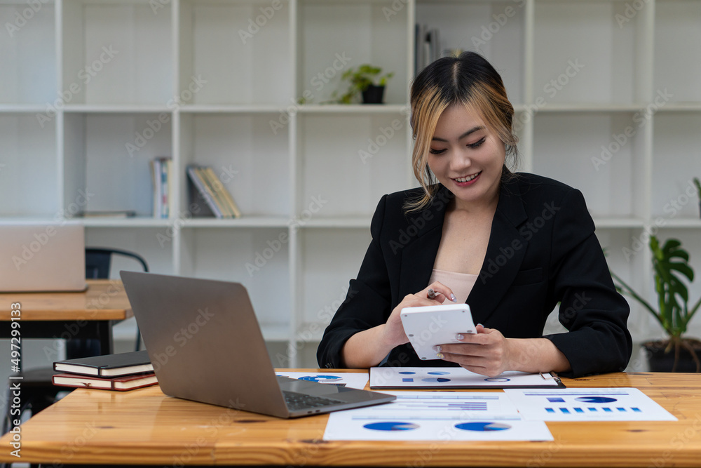 Beautiful Asian businesswoman analyzing chart using calculator, laptop at the office with papers and holding pen, sitting on the chair at modern home studio. Mobile worker contacting customers Beautiful Asian businesswoman analyzing chart using calculator, laptop at the office with papers and holding pen, sitting on the chair at modern home studio. Mobile worker contacting customers