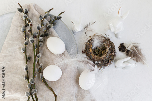 Stylish Easter flat lay. Natural easter eggs, nest, bunny, pussy willow branches, feathers, modern plate with napkin on white wooden table. Modern Easter table decoration, still life