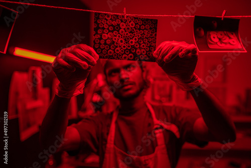 Selective focus shot of young Indian man wearing protective gloves and apron hanging wet photos on thread in darkroom