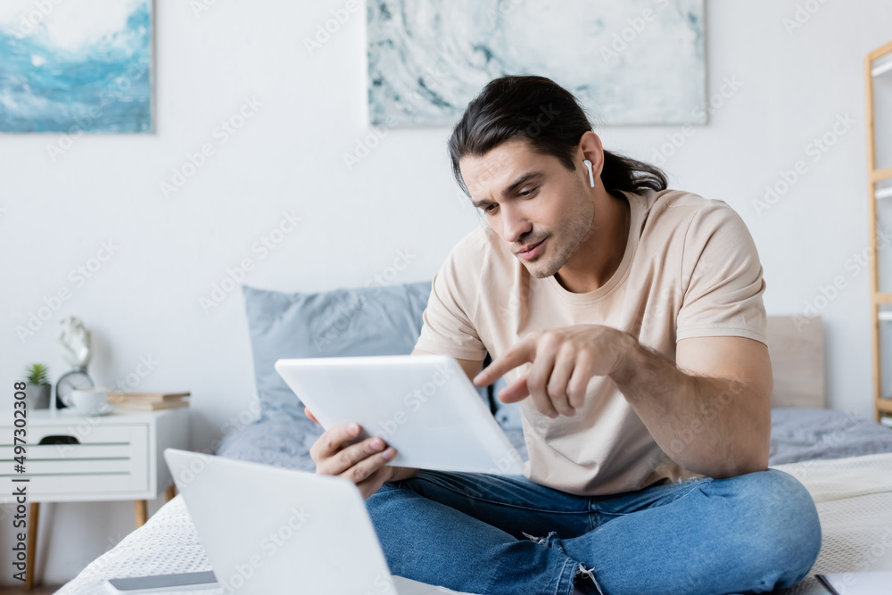 man in earphone pointing at digital tablet near laptop and smartphone on bed.