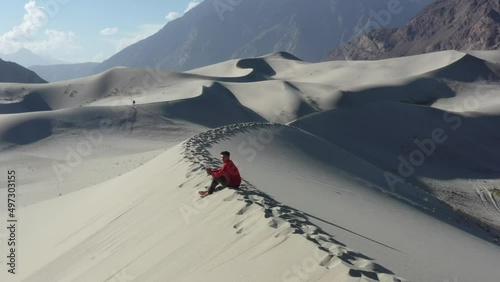 young male piloting a drone in a red jacket sitting on top of sand dunes in the cold desert of skardu pakistan during a sunny day with large mountains in the distance photo