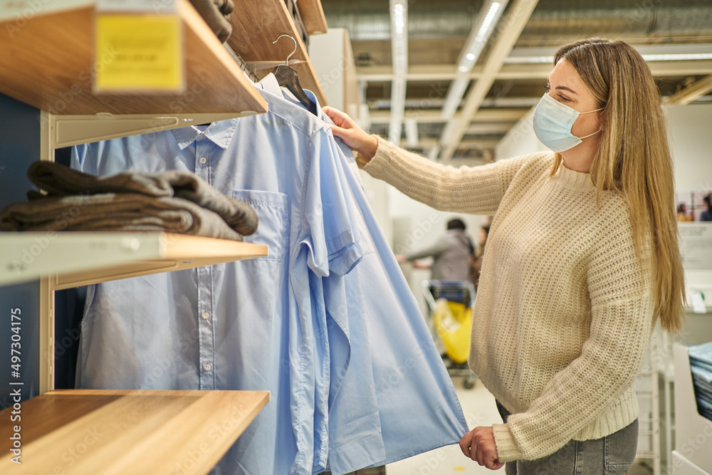 beautiful young woman in a protective mask chooses clothes in a store hung on hangers beautiful young woman in a protective mask chooses clothes in a store hung on hangers