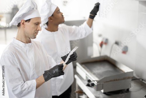 Two well-dressed chefs look on printed checks with orders while cooking in the professional kitchen. Latin-American and caucasian guys working together at restaurant