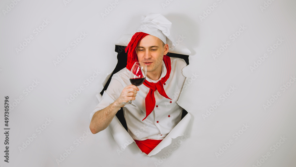Young handsome man dressed as chef with glass of wine. Male cook drinking alcoholic beverage and sticking out of hole of white background.