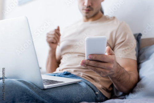 cropped view of man holding smartphone near laptop in bedroom.