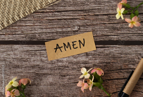 Amen, handwritten text with uppercase letters in English on a wooden table with flowers and pen in vintage style. Top view, flat lay.