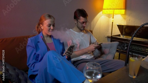 A young woman and a man are sitting in a hookah bar, smoking and drinking alcohol. A guy and a girl are looking at their phones.