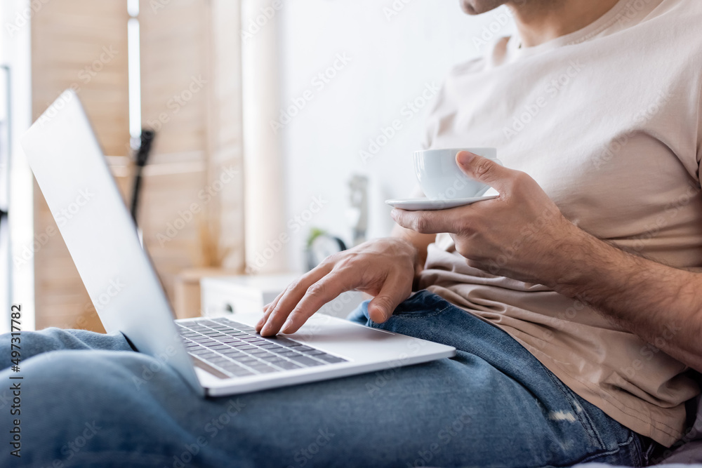 Naklejka premium cropped view of man holding cup of coffee while using laptop in bedroom.