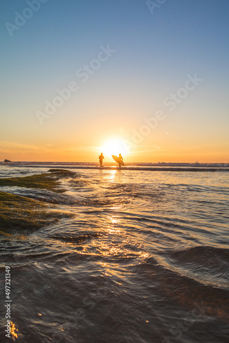 The silhouette of two surfers coming out of the ocean while the sun is setting behind them