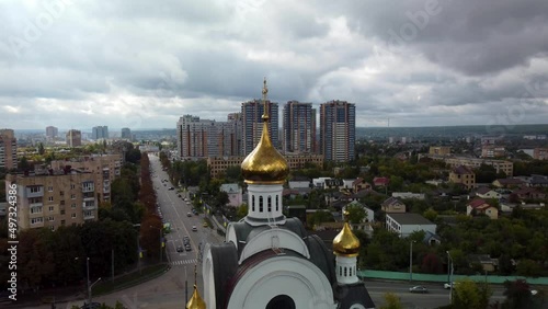 White christian church with golden dome and cross, aerial backwards flight footage with epic clouds. Kharkiv city Pavlovo Pole district, view on Nauky ave and multistory residential buildings. Ukraine photo