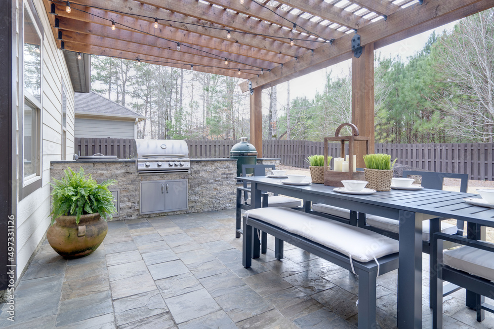 Pretty summer outdoor kitchen with table set and grilling station underneath wooden arbor on stone patio. - obrazy, fototapety, plakaty 