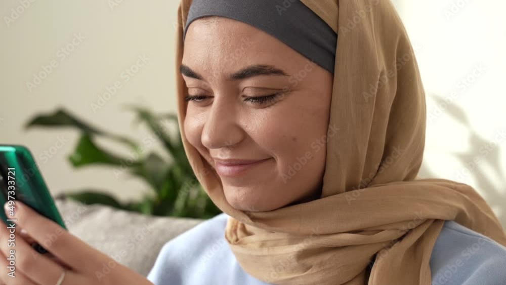 Portrait of positive calm muslim lady wearing hijab reading good news online on smartphone, typing and sitting indoors in modern apartment. Internet communication. close up. 4k footage Portrait of positive calm muslim lady wearing hijab reading good news online on smartphone, typing and sitting indoors in modern apartment. Internet communication. close up. 4k footage