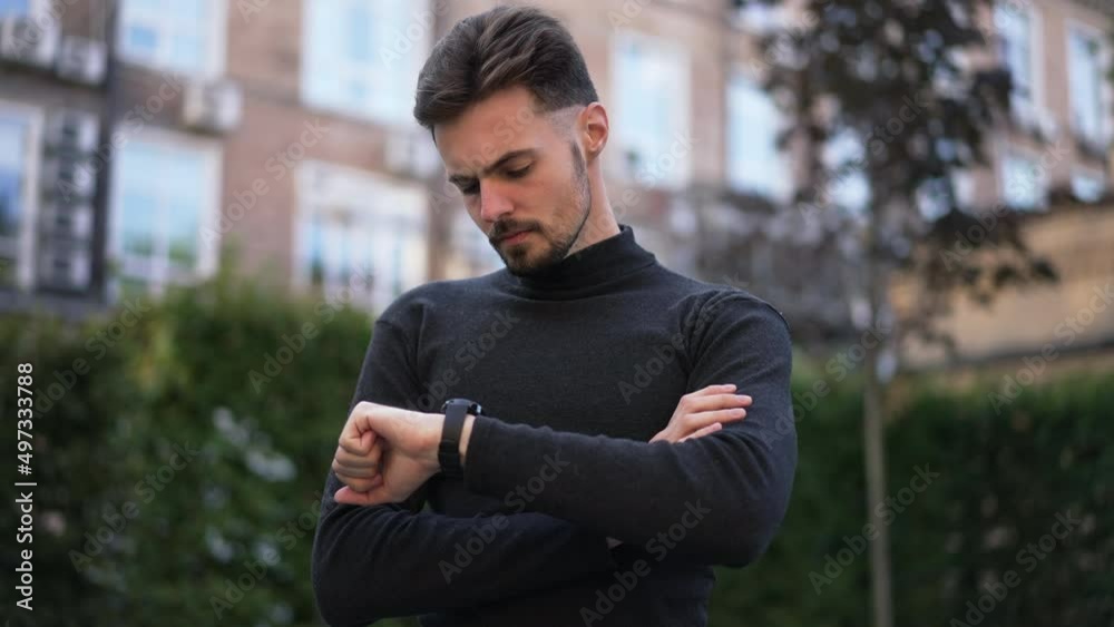 Portrait of serious Caucasian young businessman checking time on wristwatch looking around standing outdoors. Confident intelligent handsome man waiting on city street. Slow motion Portrait of serious Caucasian young businessman checking time on wristwatch looking around standing outdoors. Confident intelligent handsome man waiting on city street. Slow motion