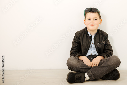 Schoolboy in a leather jacket and glasses sits on the floor. Cool boy on a white background. Copy space