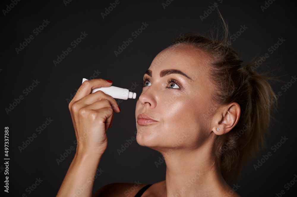 Fototapeta premium Close-up portrait of a bautiful middle aged woman applying anti puffiness anti wrinkles moisturizer under eyes. Youth concept and prevention of first signs of aging process.