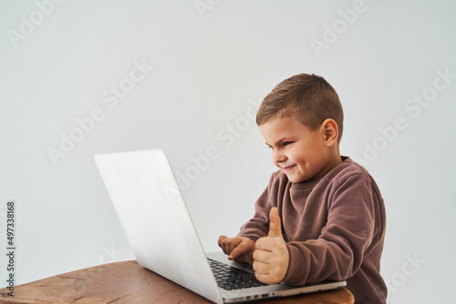 Child boy study online with laptop and shows thumbs up and smiles. Online education and e-learning. Child taking online courses on laptop.