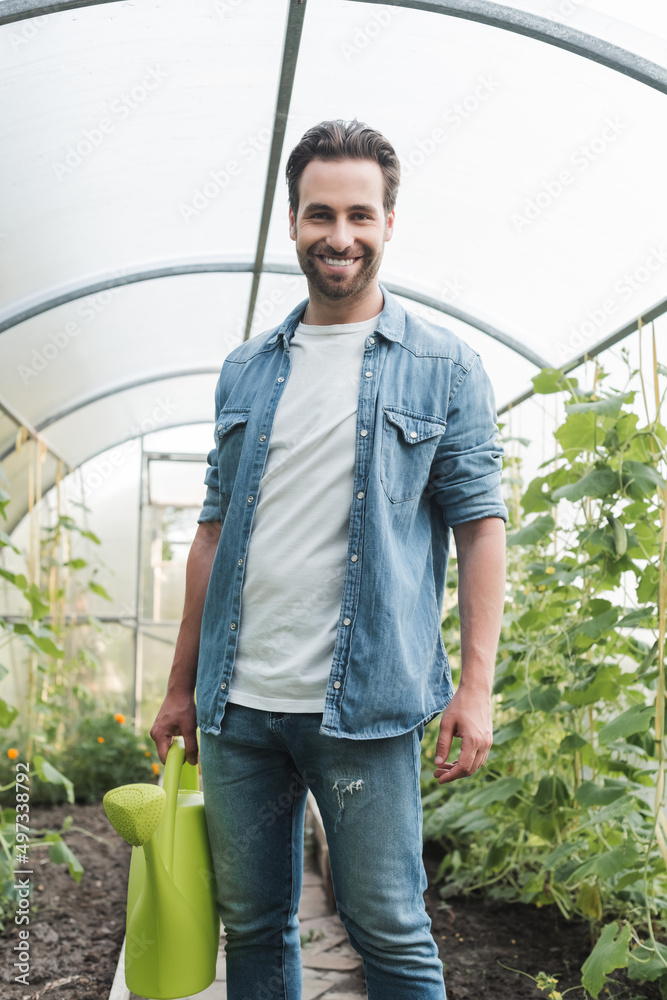 Fototapeta premium young farmer with watering can smiling at camera in greenhouse.
