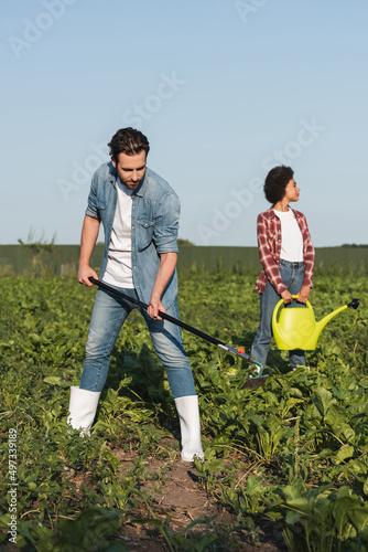 full length view of farmer working with hoe near african american woman with watering can.