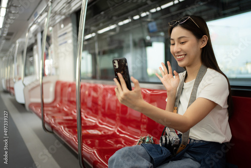 Selective focus of a smiling Asian female traveler sitting on a red row chair in an empty electric train, waving hand while video call on a cellphone during travel by sky train. Copy space on the left