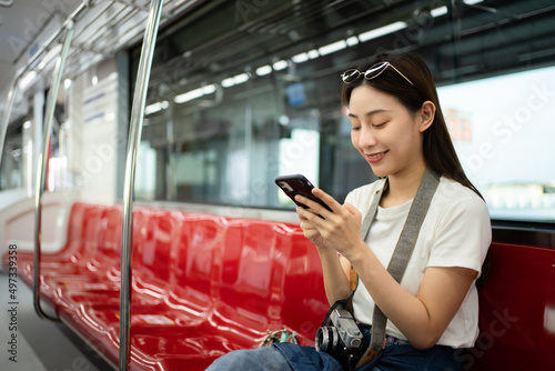 Selective focus of a happy Asian female traveler with sunglasses sitting on a red row chair with no other passengers in the electric train, smiling using a cellphone while traveling by the sky train.
