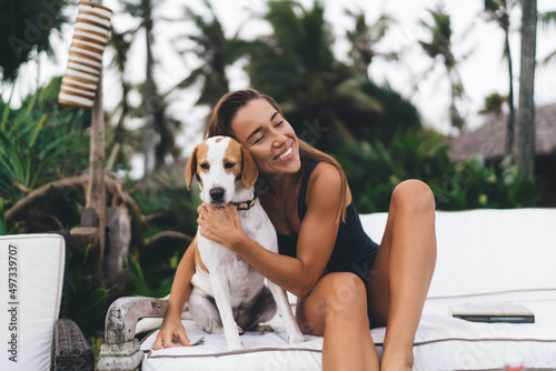 Cheerful woman in swimwear recreating at VIP lounge zone with favorite purebred dog in collar, happy Caucaisan hipster girl enjoying friendship with lovely mongrel doggie - resting at coastline