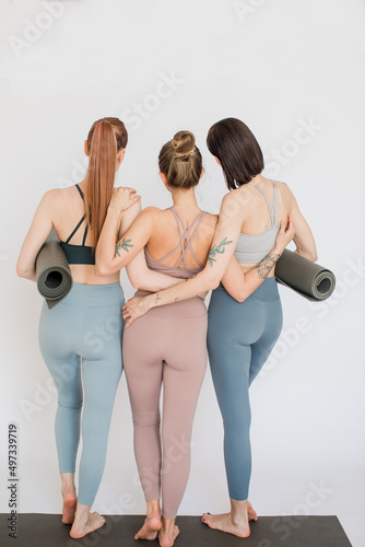 three girls in beautiful sportswear hugging each other, holding yoga mats in their hands, view from the back, long shot