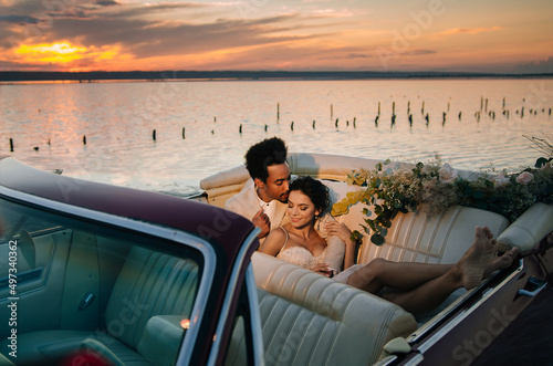 Beautiful couple at sunset on a summer wedding day. Romantic couple in love next to their convertible. The groom is in a light suit, the bride is in a beautiful stylish dress. © ann
