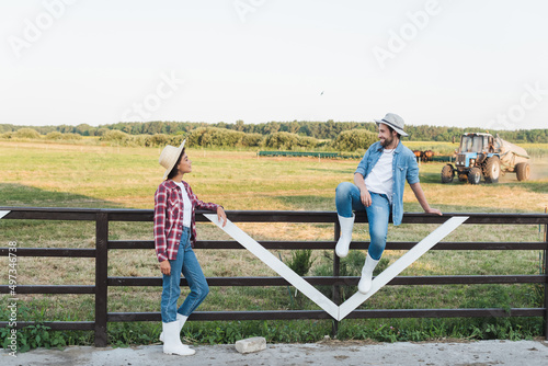 full length view of smiling farmer sitting on wooden fence near young african american colleague.