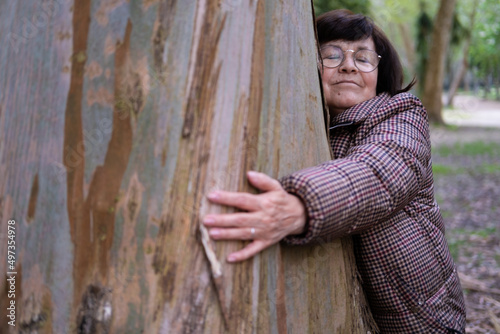 A smiley senior woman with glasses hugging a massive wide tree trunk with her eyes closed in the forests