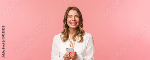 Close-up portrait of charming, lovely blond girl using mobile phone, texting friend, order in online store using smartphone application, laughing and smiling pleased, stand pink background