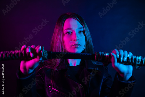Cyber girl in a black leather jacket at dusk holds a katana. A woman in a club with a colored pink-blue light holds an Asian sword. photo