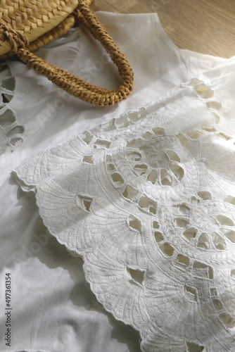 White linen lace blouse and round straw bag on wooden background. Selective focus.