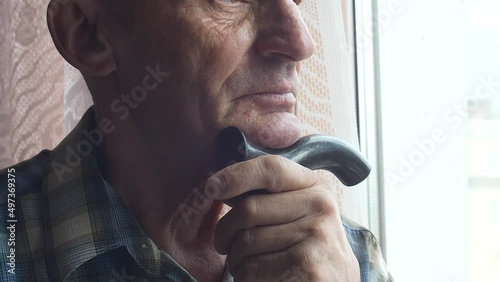 Portrait of a sad caucasian pensioner at the window waiting with a walking stick.Close-up of the wrinkled face of a lonely elderly man with a cane.Nursing home theme. Selective focus photo