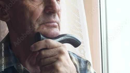 Portrait of a serious Caucasian pensioner over 70 years old in a walking stick with a cane looks out the window and waits for his relatives. Lonely old age theme. Nursing home. Elderly lonely man photo