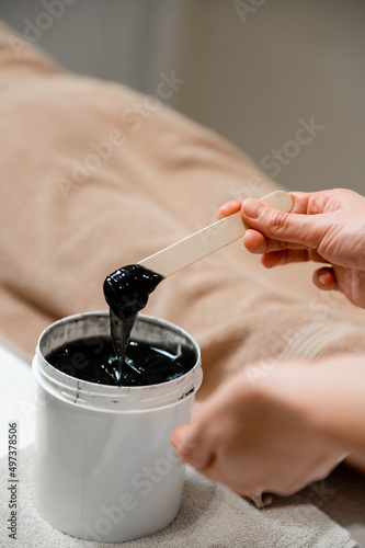 The girl holds a wooden spatula to apply a close-up of the scrub. The girl collects a black depilatory agent from a bucket close-up. Black scrub.