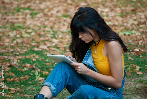 Young girl dressed in yellow and wearing denim overalls sitting while writing or drawing in a notebook, park of autumn green and yellow colors