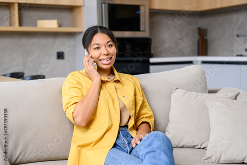 Happy African-American woman talks via smartphone sitting on the sofa at home, charming lady enjoys phone conversation staying at home, glad to talk with friend
