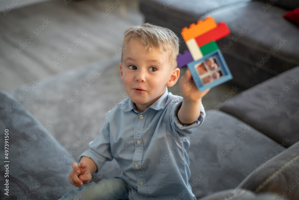 Fototapeta premium A small child plays with blocks. The boy sits on the sofa and builds a house from the constructor.