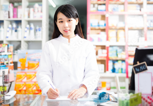 Confident chinese female pharmacist standing with a cash desk in drugstore