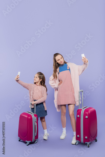 full length of happy mother and child taking selfie on mobile phones near baggage on purple.