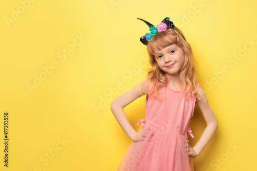 Close-up portrait of a cute attractive cheerful girl. Cute smiling child wearing a festive unicorn headband. Blonde girl 5 years old in a pink dress on a bright yellow background looking at the camera