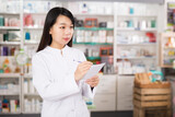 Chinese female looking medicine with notebook near shelves in pharmacy. High quality photo