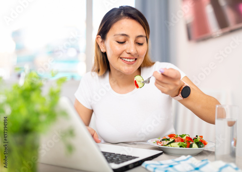Young peruvian housewoman eating vegetable salad and watching movie on laptop photo