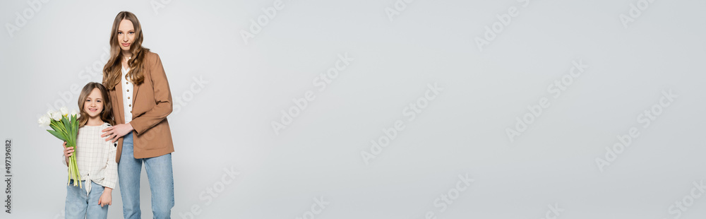 woman looking at camera near smiling daughter with tulips bouquet isolated on grey, banner. woman looking at camera near smiling daughter with tulips bouquet isolated on grey, banner.